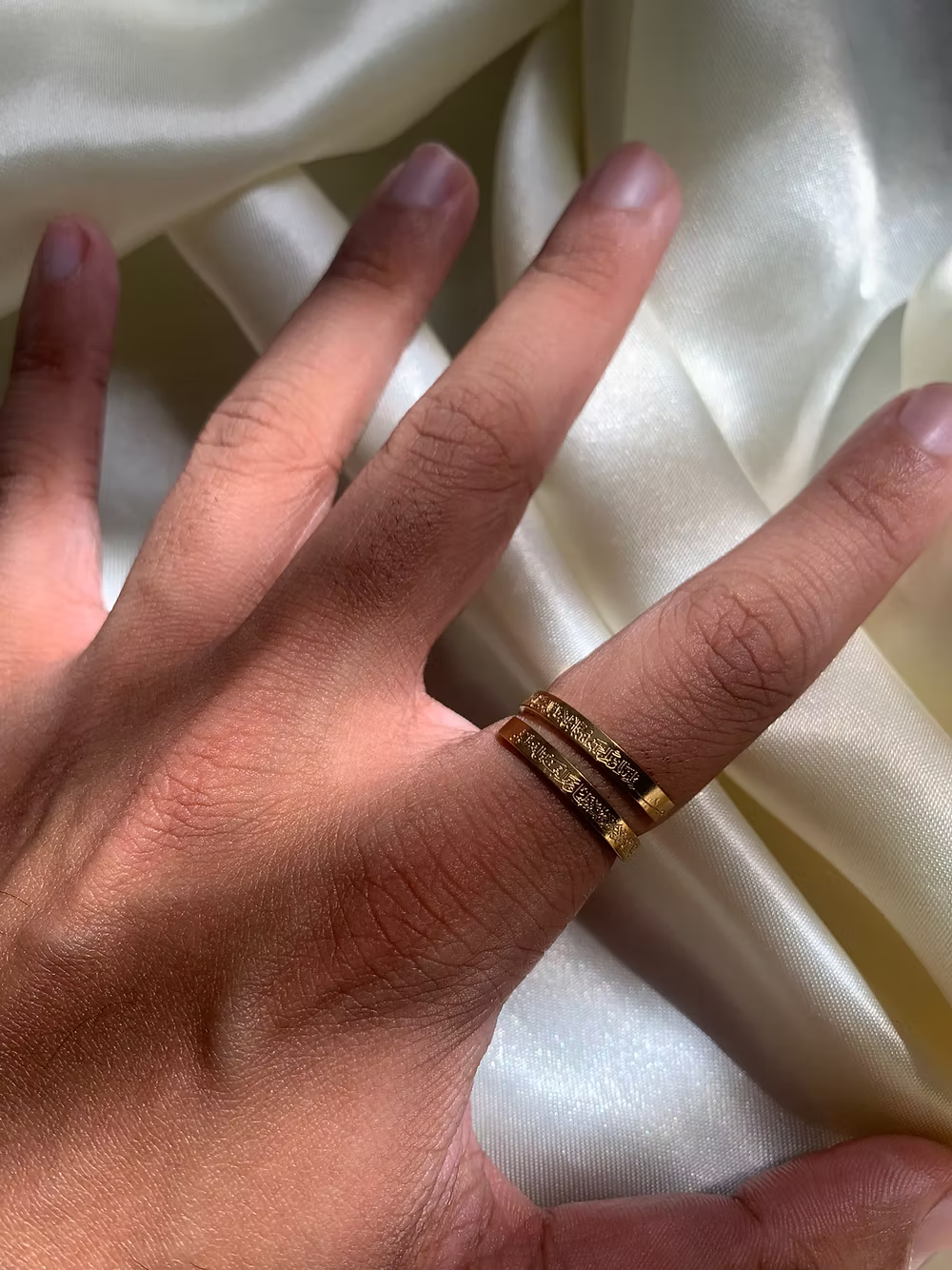 Hand wearing a gold Ayatul kursi ring with engraved text on a white satin background
