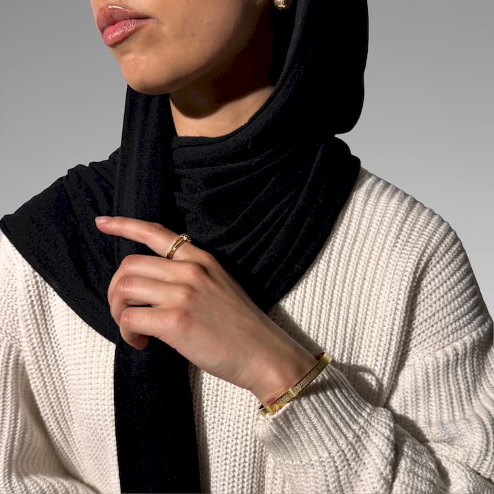 Person wearing a black hijab and beige sweater with gold jewelry on a neutral background