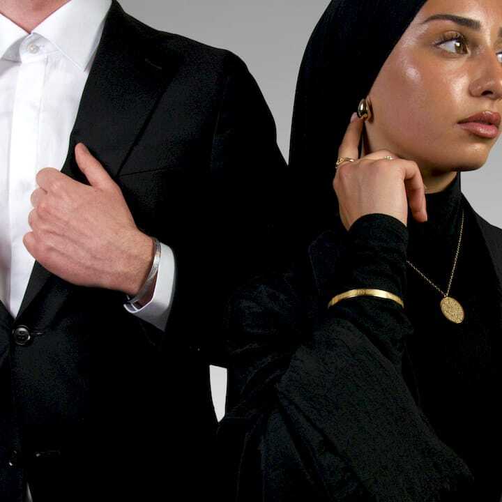 Man in a suit and woman in a black outfit with gold islamic jewelry against a gray background